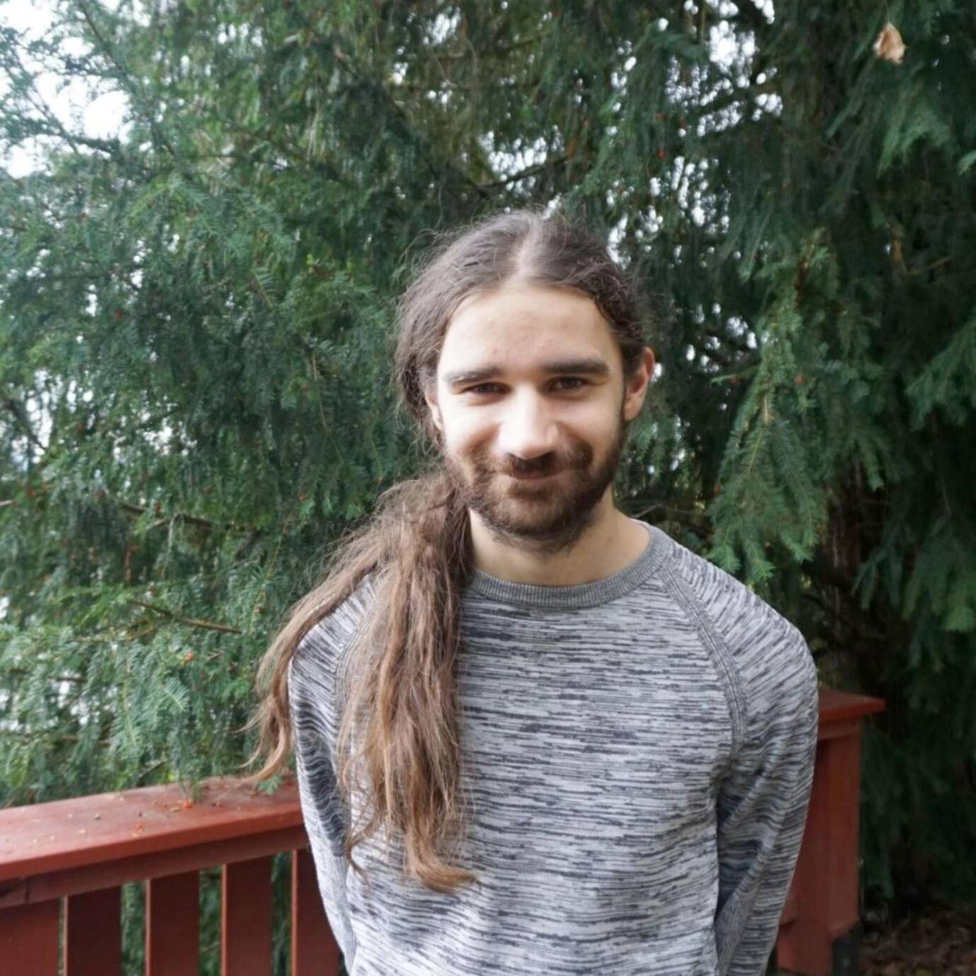 Eine lächelnde Person steht vor einem Zaun und Nadelbäumen