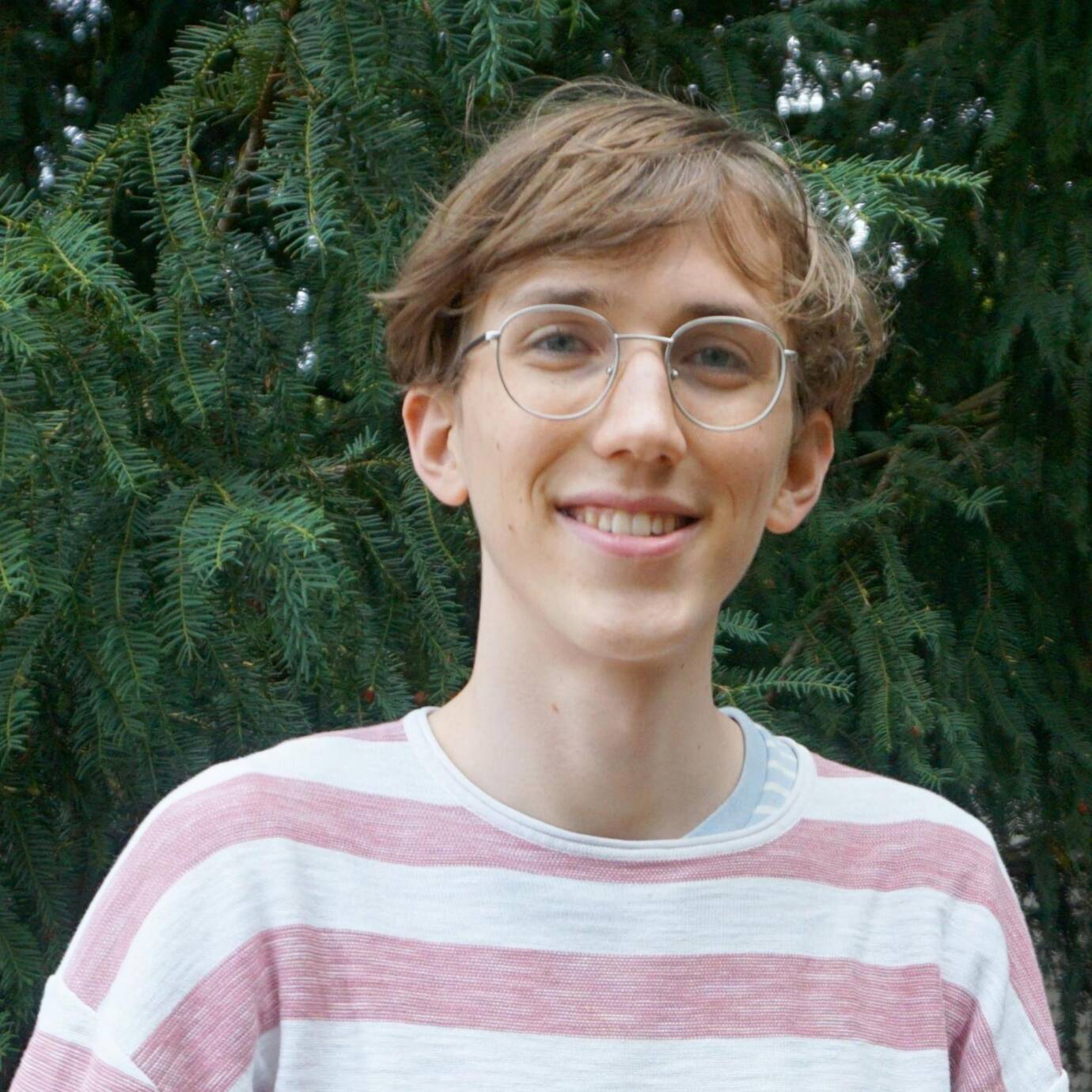 Eine lächelnde Person steht vor Nadelbäumen