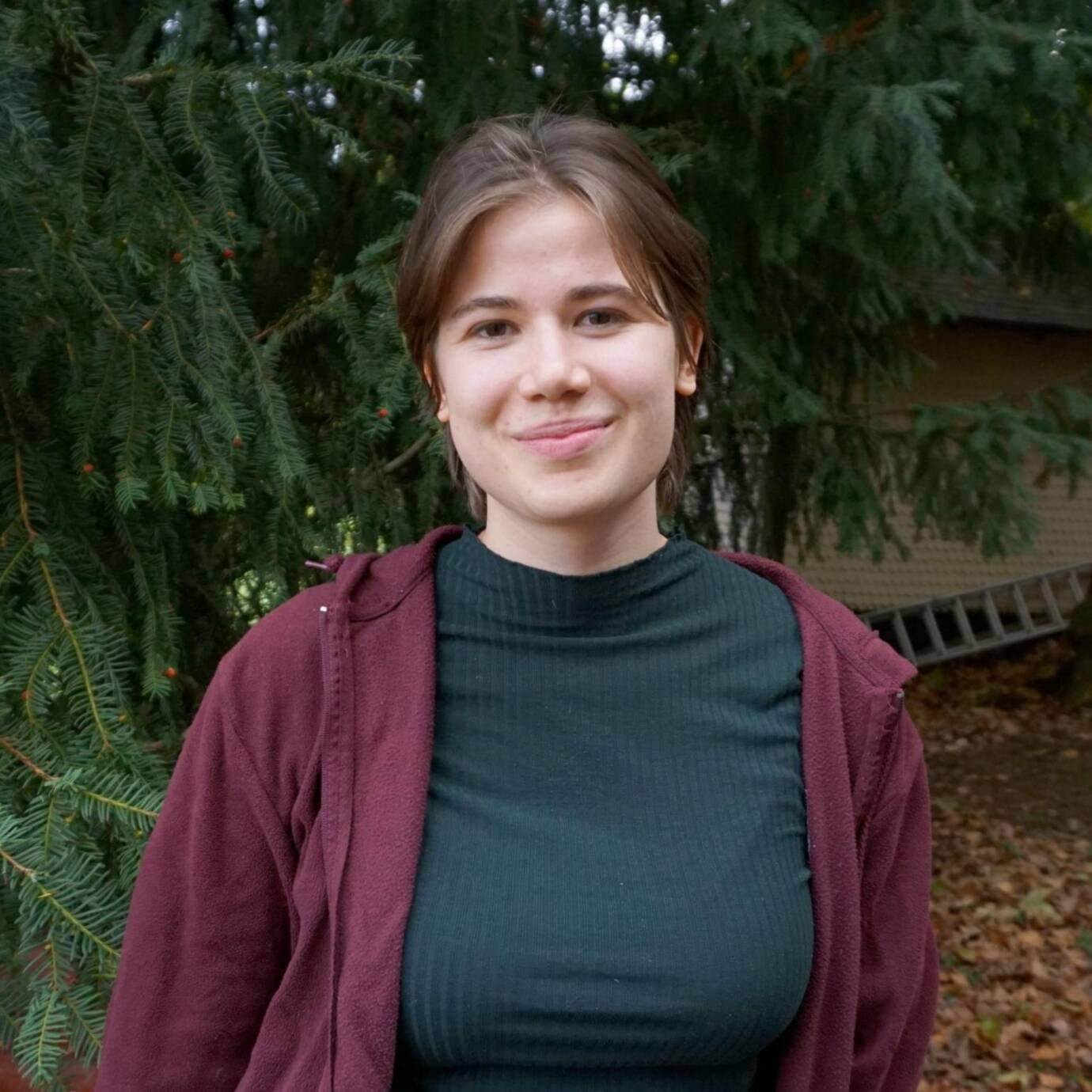 Eine lächellnde Person steht vor Nadelbäumen