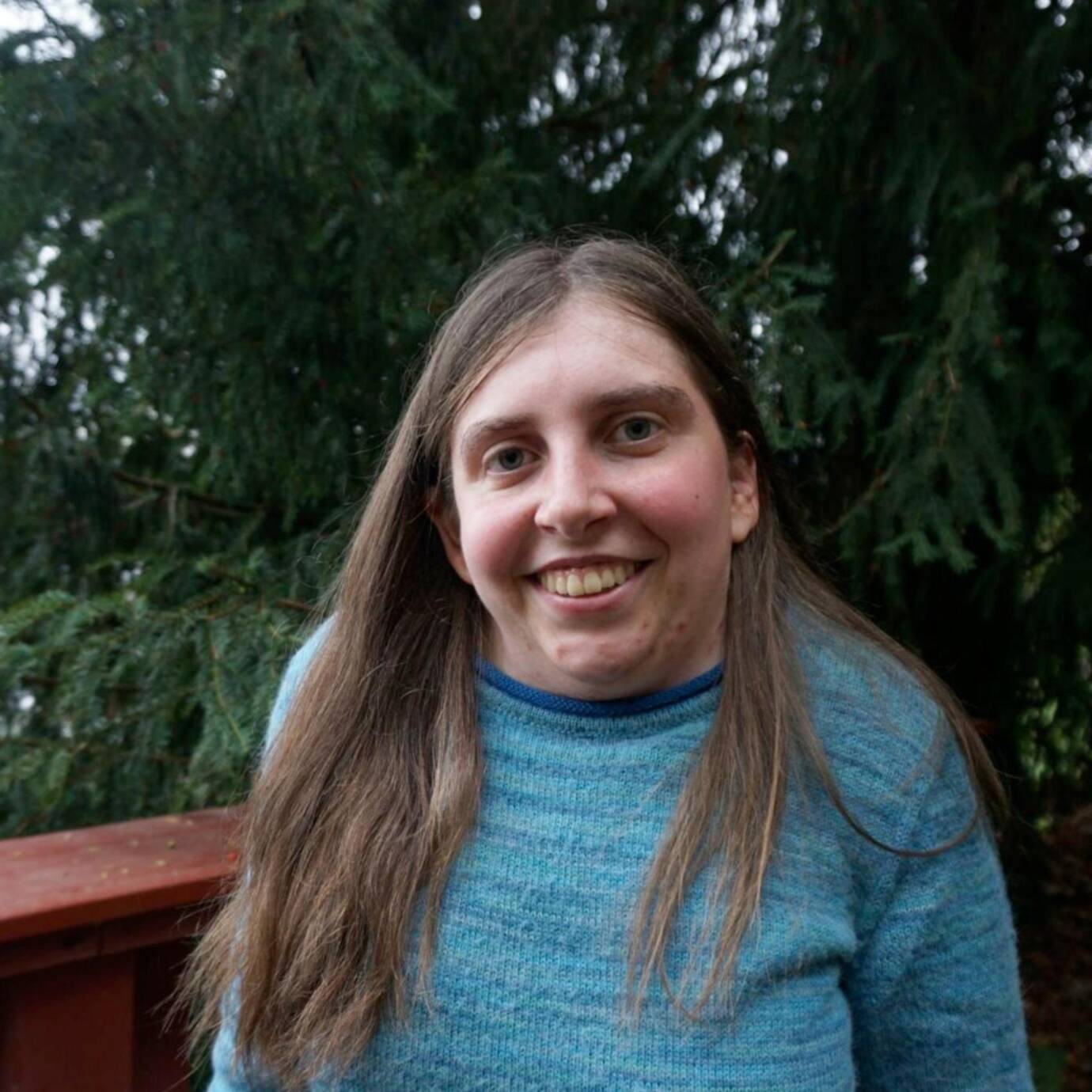 Eine lächelnde Person steht vor einem Zaun und Nadelbäumen