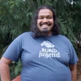 Eine lächelnde Person, im blauen BUNDjugend-T-shirt steht vor Nadelbäumen