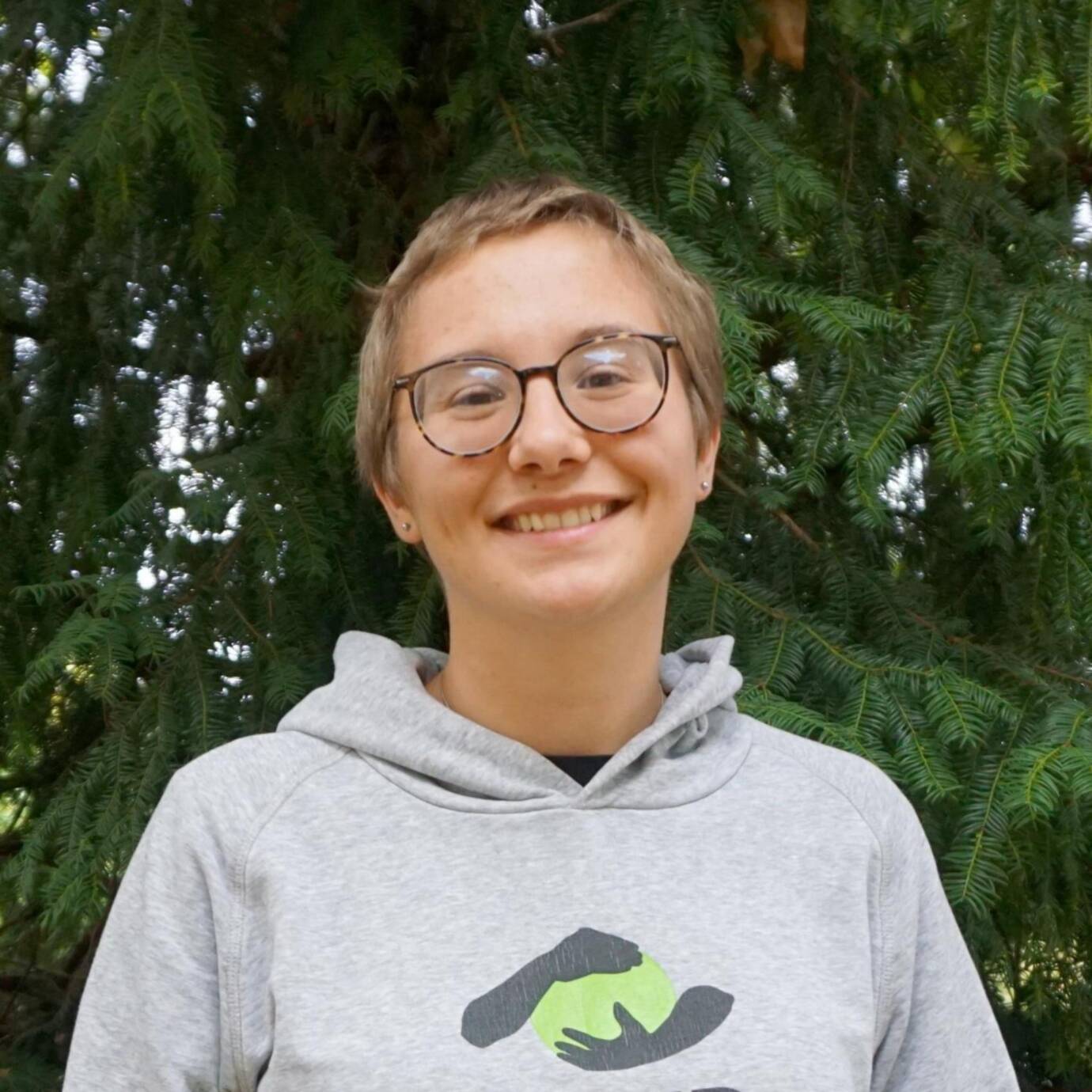 Eine lächelnde Person mit BUNDjugend-Hoodie steht vor Nadelbäumen